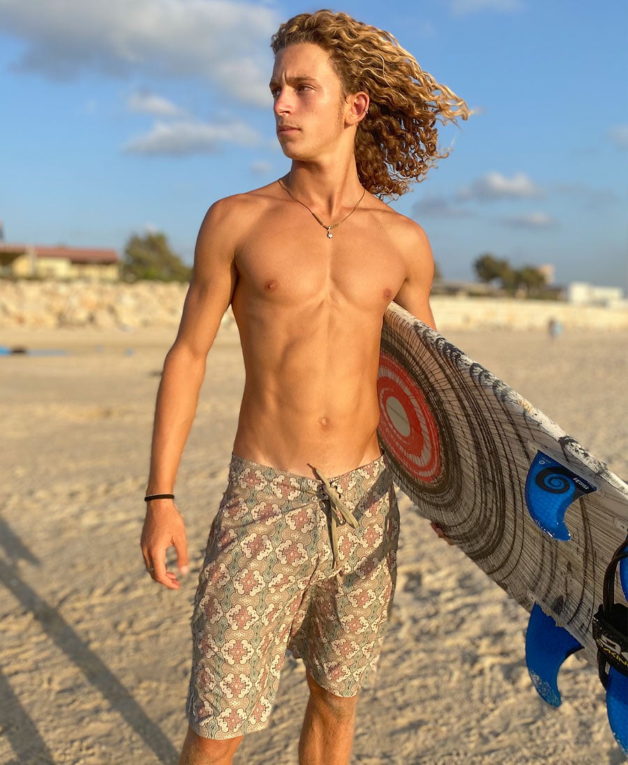 Man in Shipibo print boardshorts holding a surfboard on the beach